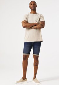 Camiseta beige clara, pantalones cortos de mezclilla oscuros con dobladillos enrollados y zapatos tipo mocasín beige. Presenta tatuajes visibles en los brazos y las piernas.
