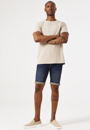 Lichte beige t-shirt, donkere denim shorts met opgerolde zoom, en beige instappers. Heeft zichtbare tatoeages op de armen en benen.