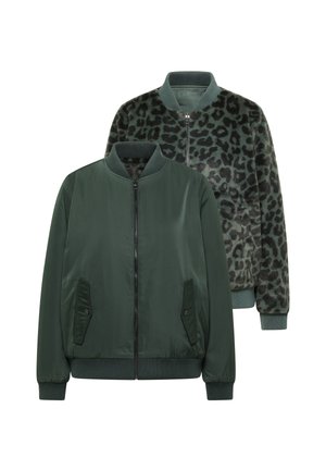 Femme portant une veste zippée verte, un jean large foncé, des baskets grises et une casquette jaune pâle, debout contre un mur beige bicolore.