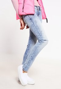 Vaaleansiniset farkut, joissa on kapea malli, haalistunut pinta ja hieman kulutusta, yhdistettynä valkoisiin liukuhyppytossuihin ja vaaleanpunaiseen puffereeseen.