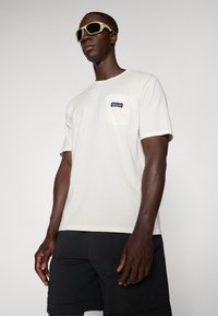 Light cream short-sleeve t-shirt with a pocket on the left chest featuring a blue label. Worn with dark shorts and sunglasses.