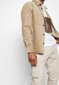 Beige ribfluwelen overhemd met knopen en een borstzak, gedragen over een wit T-shirt, gecombineerd met lichtbeige cargobroek en een bruine crossbodytas.