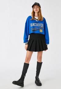 Skórzany niebieski sweter z logo "Vancouver", czarna spódnica plisowana oraz czarne skórzane kozaki do kolan z grubą podeszwą. Czapka w kolorze czarnym jako dodatek.
