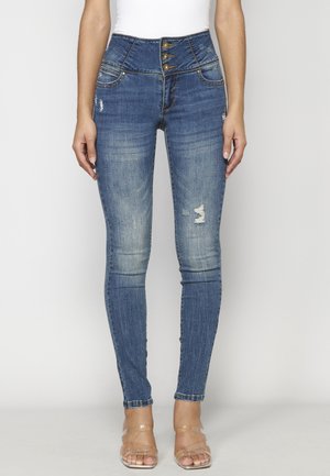 Femme portant un jean skinny bleu déchiré taille haute et des talons ouverts avec brides transparentes, se tenant devant un fond clair uni.