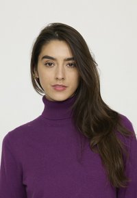 Pull à col roulé violet, texture côtelée, coupe ajustée et manches longues. Le mannequin a les cheveux longs et porte peu d'accessoires.