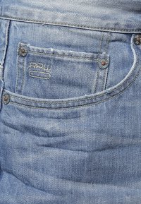 Jeans de algodón de mezclilla, de color azul claro, con una textura suave. Presenta un bolsillo trasero con remaches de metal y un detalle de logo cosido.