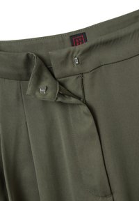Gros plan de la taille d'un pantalon vert olive montrant un fermoir en métal à crochet et œillet, avec une étiquette de vêtements noire et rouge partiellement visible.