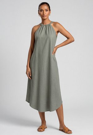 Kobieta stojąca w zielonej oliwkowej sukience midi bez rękawów z marszczonym dekoltem, nosząca beżowe płaskie sandały, na tle jednolitego szarego tła.