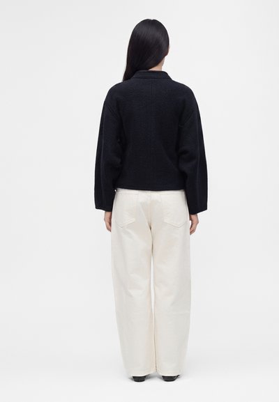 Femme aux longs cheveux foncés, vue de dos, portant un haut noir à manches longues et un pantalon blanc à jambes larges sur un fond uni.