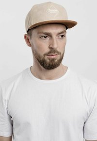 Tan cap with a brown brim, featuring a white logo on the front. Worn with a plain white t-shirt. The texture appears soft and smooth.