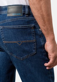 Dunkelblaue Denim-Jeans mit einer Gesäßtasche, die ein stilisiertes Nahtdesign und ein schwarzes Etikett mit "pierre cardin" in blauer Schrift aufweist.