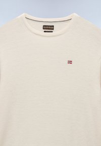 Suéter color crema hecho de un tejido suave, con cuello redondo y un pequeño logotipo de bandera bordado en el pecho. Textura liviana.