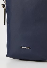 Borsa in tessuto blu navy con finitura testurizzata, dotata di una zip argentata e di un logo metallico Calvin Klein sul davanti.