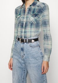 Chemise à carreaux verts et bleus à manches longues, avec deux poches poitrine et fermeture par boutons, associée à un jean bleu clair taille haute et une ceinture noire.