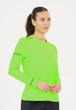 Vrouw met donker haar in een paardenstaart, die een felgroen shirt met lange mouwen en zwarte broek draagt, staand met de handen voor zich samengevouwen.