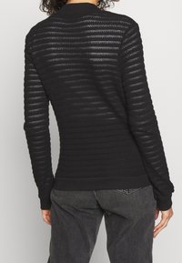 Person wearing a black, long-sleeve, semi-sheer knit sweater with horizontal stripes and dark gray jeans, shown from the back.