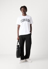 Hvit bomull T-skjorte med "CALVIN KLEIN" trykt i svart buet skrift, kombinert med svarte bukser og hvite sneakers. Modellen holder en svart totebag.