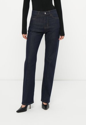 Jeans a vita alta di colore blu scuro in denim. Presentano gambe dritte, tasche anteriori e bottoni metallici argentati. Indossati con tacchi neri.