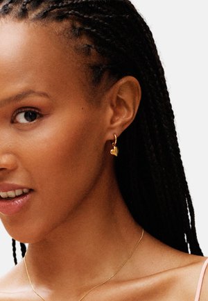 Kullast südamekujuline rõngaskõrvarõngas läikiva viimistlusega, ripub väikese rõnga küljes, taustal põimitud juuksed ja õrn roosa top.