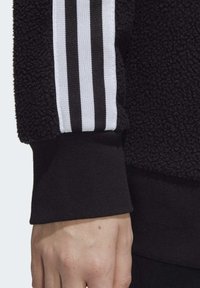 Svart fleecetröja med en texturerad tyg, vita och svarta randiga detaljer på ärmen och en ribbad mudd.