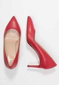 Une paire de chaussures à talons hauts rouges à bout pointu, une chaussure montrant une vue de dessus et l'autre une vue de côté sur un fond blanc.