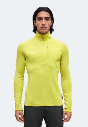 Homme aux cheveux foncés porte un haut vert citron à manches longues avec une fermeture éclair quart de zip et une poche poitrine, ainsi qu’un pantalon noir, debout devant un fond uni.