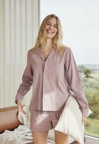 Vaaleanlila pyjamapuku, jossa on napitettava pitkähihainen paita, jossa on tasku, ja siihen sopivat shortsit. Materiaali on teksturoitu ja malli on rento.
