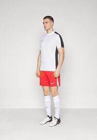 Witte sportshirt met korte mouwen en zwarte schouderaccenten, gecombineerd met rode shorts met witte zijstrepen en een Nike-logo, aangevuld met witte sokken en zwarte voetbalschoenen.