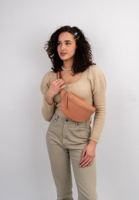 Borsa a tracolla in pelle color tan con forma curvata e dettagli in cerniera dorata, indossata sopra un maglione beige e pantaloni a vita alta color beige chiaro.