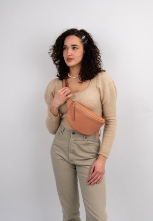Brun læder crossbody taske med en buet form og guld lynlåsdetalje, båret over en beige sweater og lys beige højtaljede bukser.