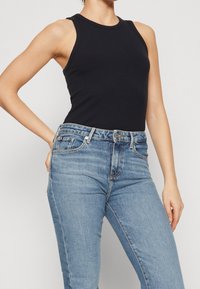 Persona che indossa un top a righe nero senza maniche infilato in jeans di denim azzurri a vita alta, in piedi con una mano sui fianchi.
