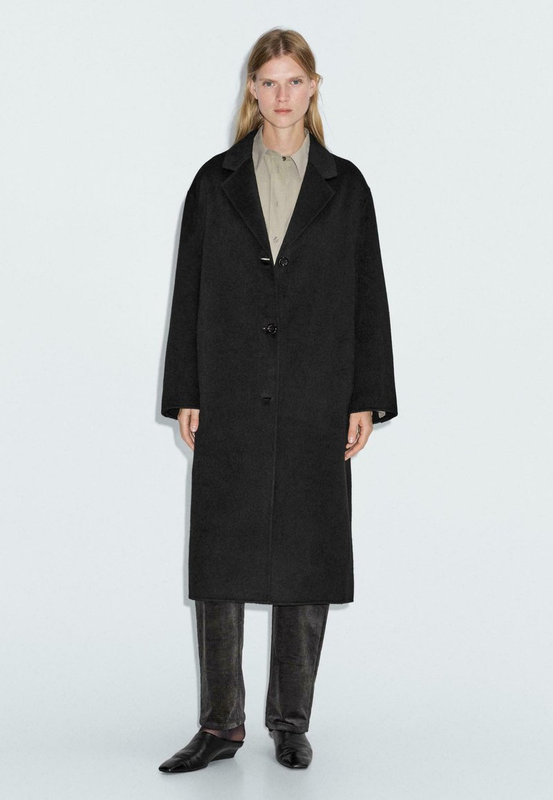 Lungo cappotto nero realizzato in tessuto testurizzato, con colletto a coste, due bottoni e maniche larghe. Indossato sopra una camicia chiara e pantaloni scuri.