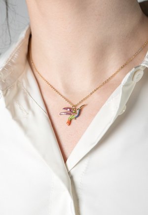 Auksinė grandinėlė su spalvingu kolibrio pakabuku, puošta violetiniais, žaliais ir oranžiniais akcentais, padėta ant baltos marškinių apykaklės.