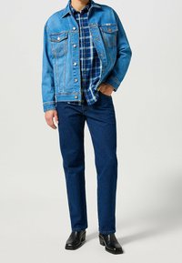 Giacca di jeans blu con bottoni metallici, abbinata a una camicia a quadri blu e verde sotto, jeans blu scuro e stivali neri alla caviglia.