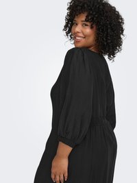 Musta mekko, jossa on rento istuvuus, muhkeat hihat ja kirjailtu vyötärö. Kangas on teksturoitu ja siluetti virtaava.