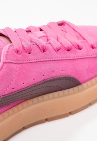 Gros plan d'une basket en daim rose avec des lacets assortis, un détail latéral marron foncé et une semelle épaisse en caoutchouc gomme.