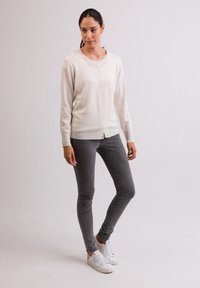 Krämfärgad kofta med knappar, rund halsringning och långa ärmar, tillsammans med grå slim fit jeans och vita sneakers. Slät textur.
