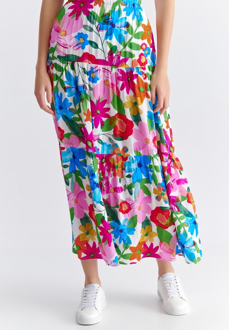 Femme portant une jupe longue fleurie colorée avec des fleurs roses, bleues, rouges et vertes, associée à des baskets blanches sur un fond uni.