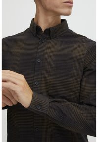 Dark plaid button-up shirt made of textured fabric, featuring a collar, black buttons, and a chest pocket. Patterns in dark brown and black.