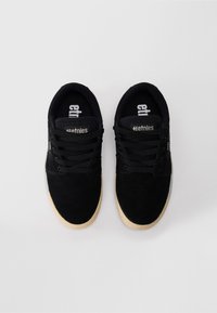 Um par de sapatos de skate etnies pretos com solas bejes e cordões pretos, exibidos de uma vista de cima sobre um fundo branco.