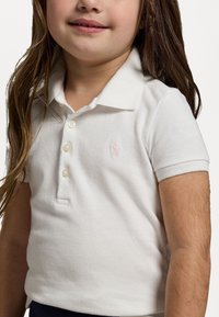 Weißes Baumwoll-Polohemd mit kurzen Ärmeln, drei Knöpfen und einem kleinen rosa gestickten Logo auf der Brust. Texturiert.