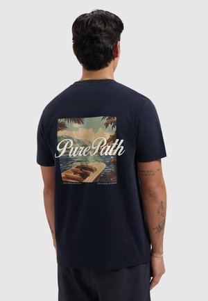 Man draagt donkerblauw T-shirt met een tropisch boottafereel en de tekst "Pure Path" op de achterkant, staand tegen een effen lichte achtergrond.