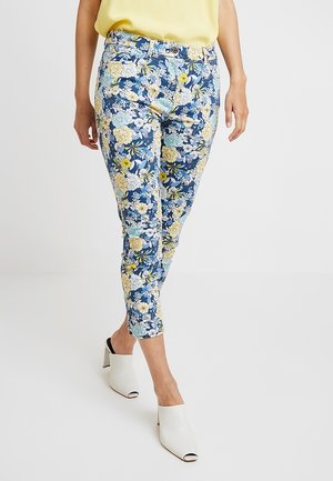 Personne portant un pantalon court bleu à fleurs, un haut jaune et des mules blanches à talons ouverts, marchant sur un fond blanc uni.