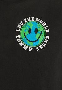 Camiseta negra con un gráfico circular de la Tierra con una cara sonriente, el texto "AMA EL MUNDO" y "TOMMY JEANS" en una fuente blanca y juguetona.