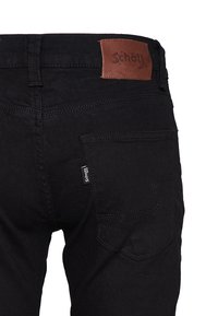 Vaqueros de mezclilla negra con un parche de cuero marrón que muestra "Schott NYC". Incluyen bolsillos traseros y un patrón de costura distintivo.