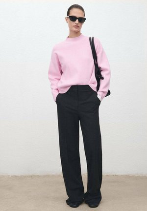 Maglione rosa a maglia con colletto alto, abbinato a pantaloni neri a vita alta e a gamba larga. Gli accessori includono occhiali da sole neri e una borsa morbida.
