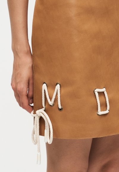 Φούστα από δέρμα χρώματος ταμπά με λευκό σκοινί περασμένο μέσα από μεταλλικά οπές σχηματίζοντας σχέδια και έναν δεμένο κόμπο κοντά στο τελείωμα, εμφανίζεται με το χέρι στο πλάι.