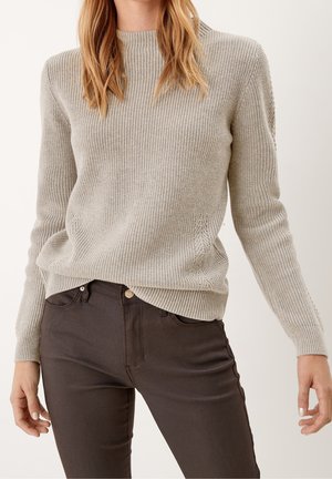 Femme portant un pull en maille beige clair à manches longues et un pantalon ajusté brun foncé, debout devant un fond uni.
