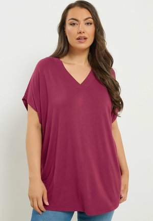 Femme aux longs cheveux bruns portant un haut magenta à manches courtes, col en V et ample, ainsi qu'un jean bleu clair, debout devant un fond blanc.