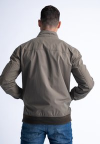 Khaki bombarďák s límečkem, žebrovanými manžetami a pasem. Hladký povrch, jednoduchý design, nošený přes modré džíny, pohled zezadu.